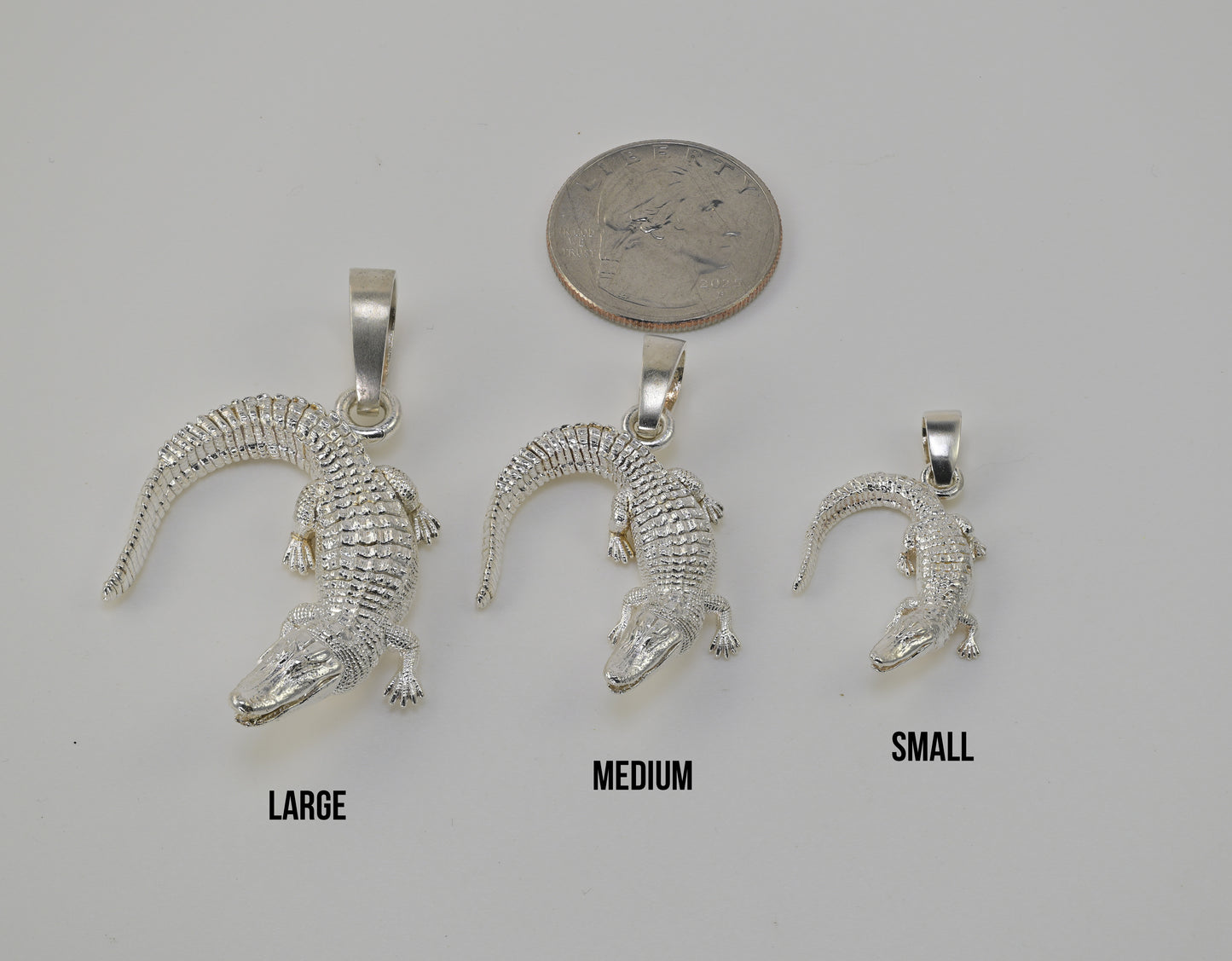 Three silver alligator pendants in large, medium, and small sizes are displayed on a light gray background with a quarter coin above them for scale and size labels 'LARGE', 'MEDIUM', and 'SMALL' underneath.