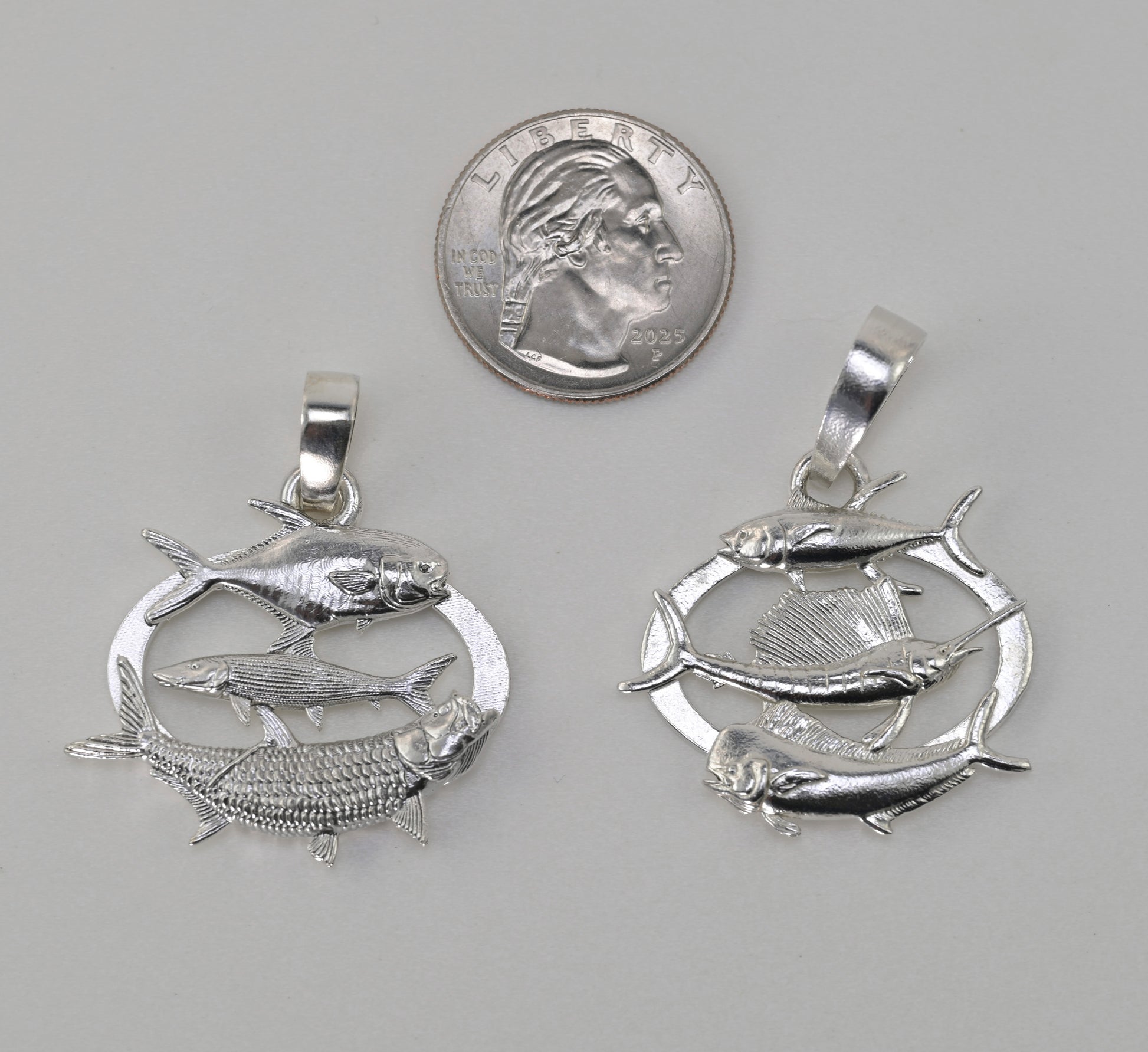 Two silver fish pendants displayed side-by-side next to a US quarter coin for scale, on a neutral gray background.
