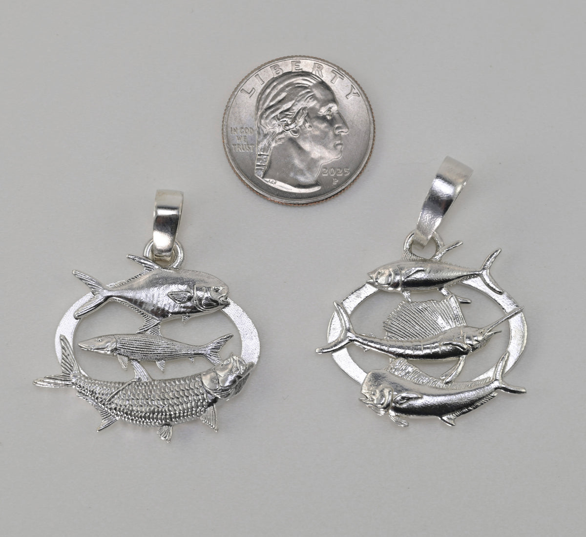 Two silver fish pendants displayed side-by-side next to a US quarter coin for scale, on a neutral gray background.