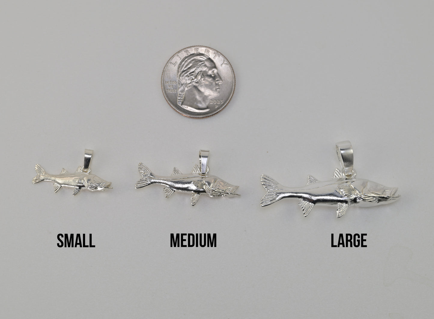 Three silvers snook fish shaped pendant charms labeled SMALL, MEDIUM, and LARGE are shown on a light gray background with a U.S. quarter