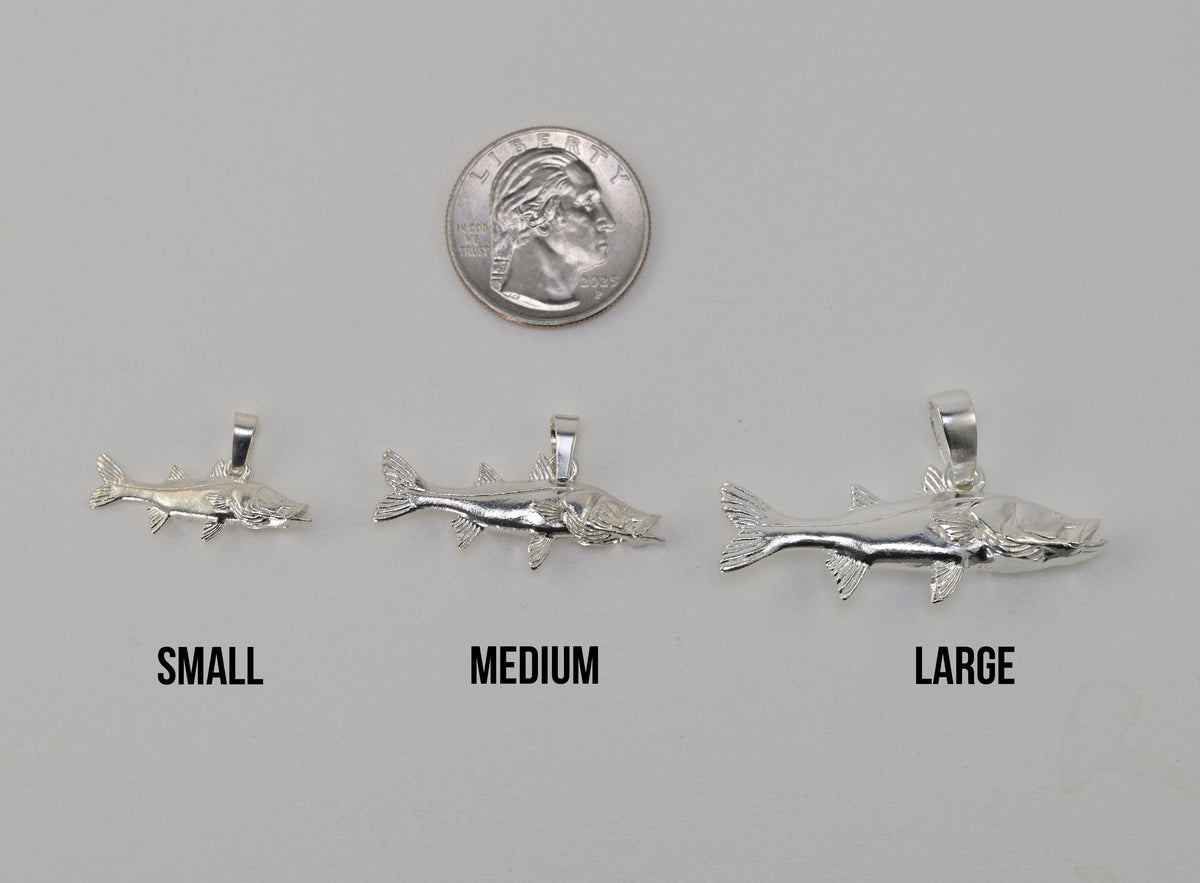 Three silvers snook fish shaped pendant charms labeled SMALL, MEDIUM, and LARGE are shown on a light gray background with a U.S. quarter